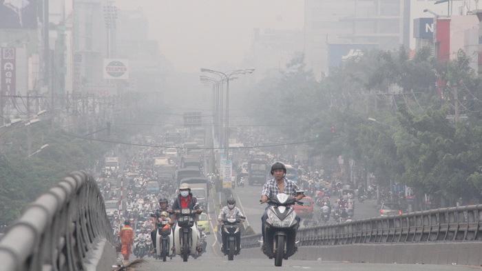 Hình ảnh cho thấy một con đường đông đúc với nhiều xe máy và ô tô di chuyển trong bầu không khí ô nhiễm nặng. Lớp khói bụi mờ bao phủ toàn bộ khung cảnh, thể hiện rõ tình trạng bụi mịn và ô nhiễm không khí nghiêm trọng ở khu vực đô thị.