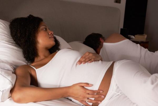 Một người phụ nữ mang thai nằm trên giường, với biểu cảm lo lắng, trong khi người đàn ông nằm cạnh không chú ý. Hình ảnh phản ánh những thay đổi hormone và sinh lý ở phụ nữ trong giai đoạn mang thai, liên quan đến rối loạn giấc ngủ.
